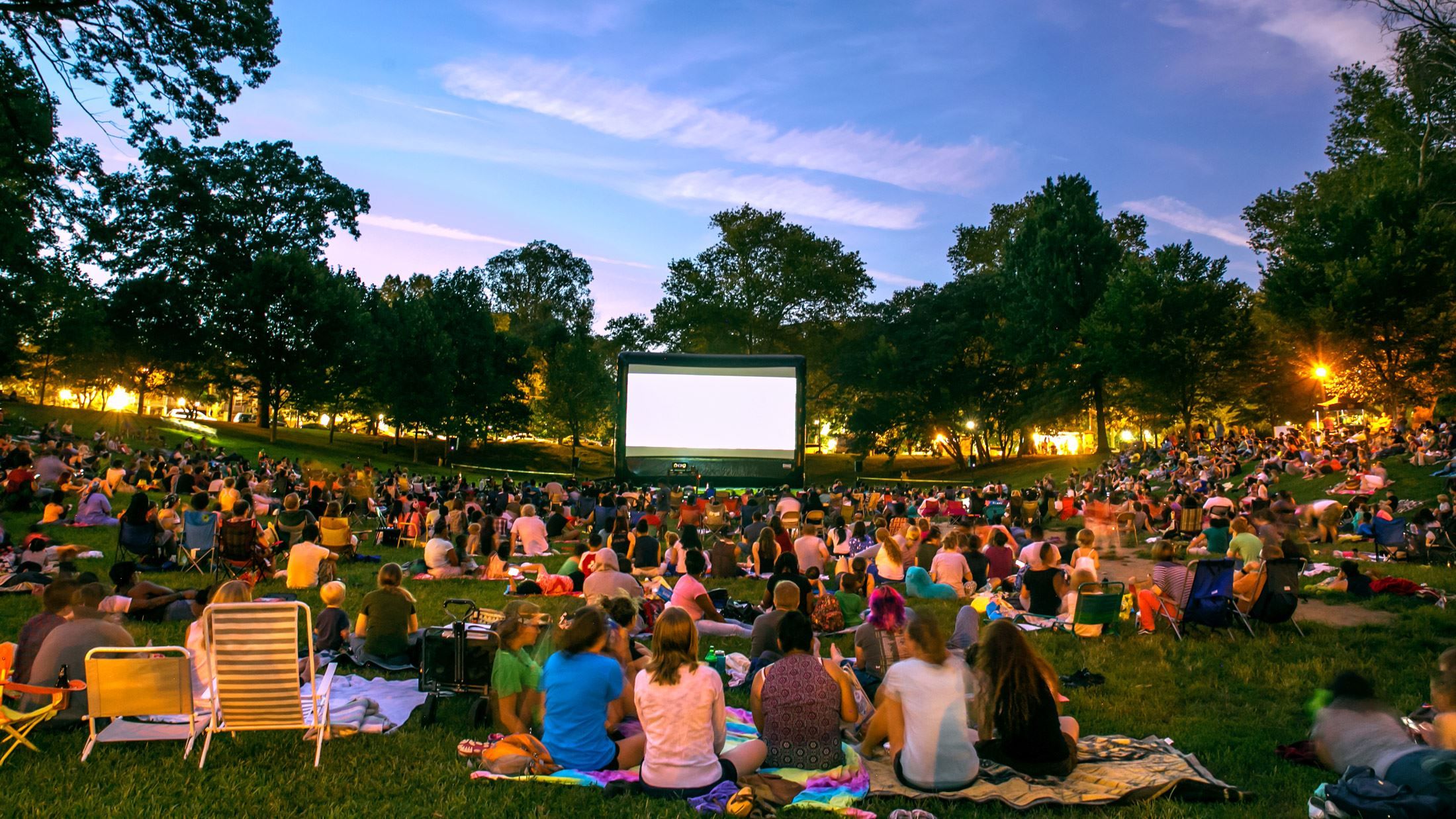 A crowd seated on blankets and lawn chairs watching an outdoor movie screen at dusk during the Goddard Fall Festival