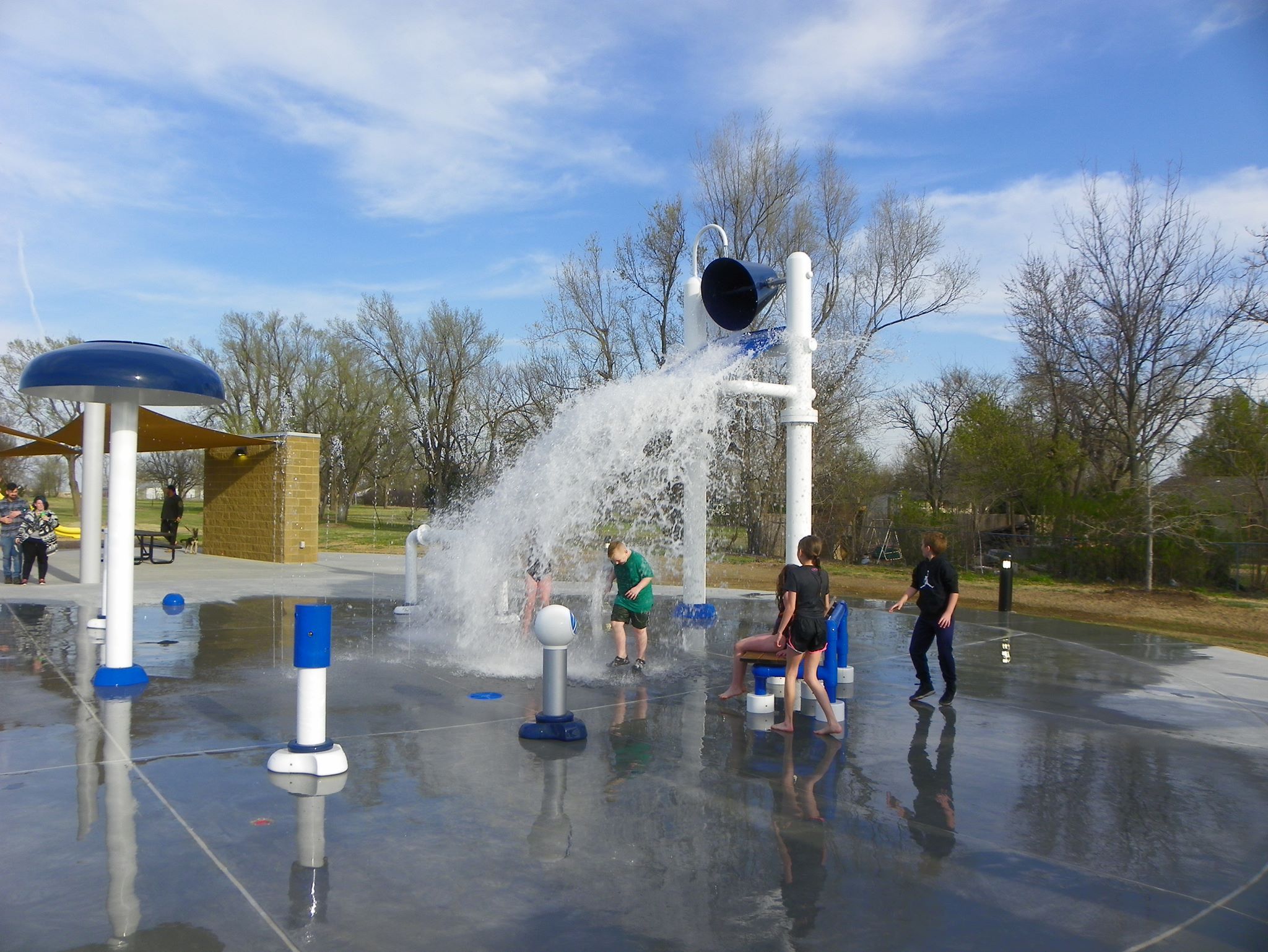 Water features running at the Goddard Linear Park splash pad, with a shade canopy and playground beyond