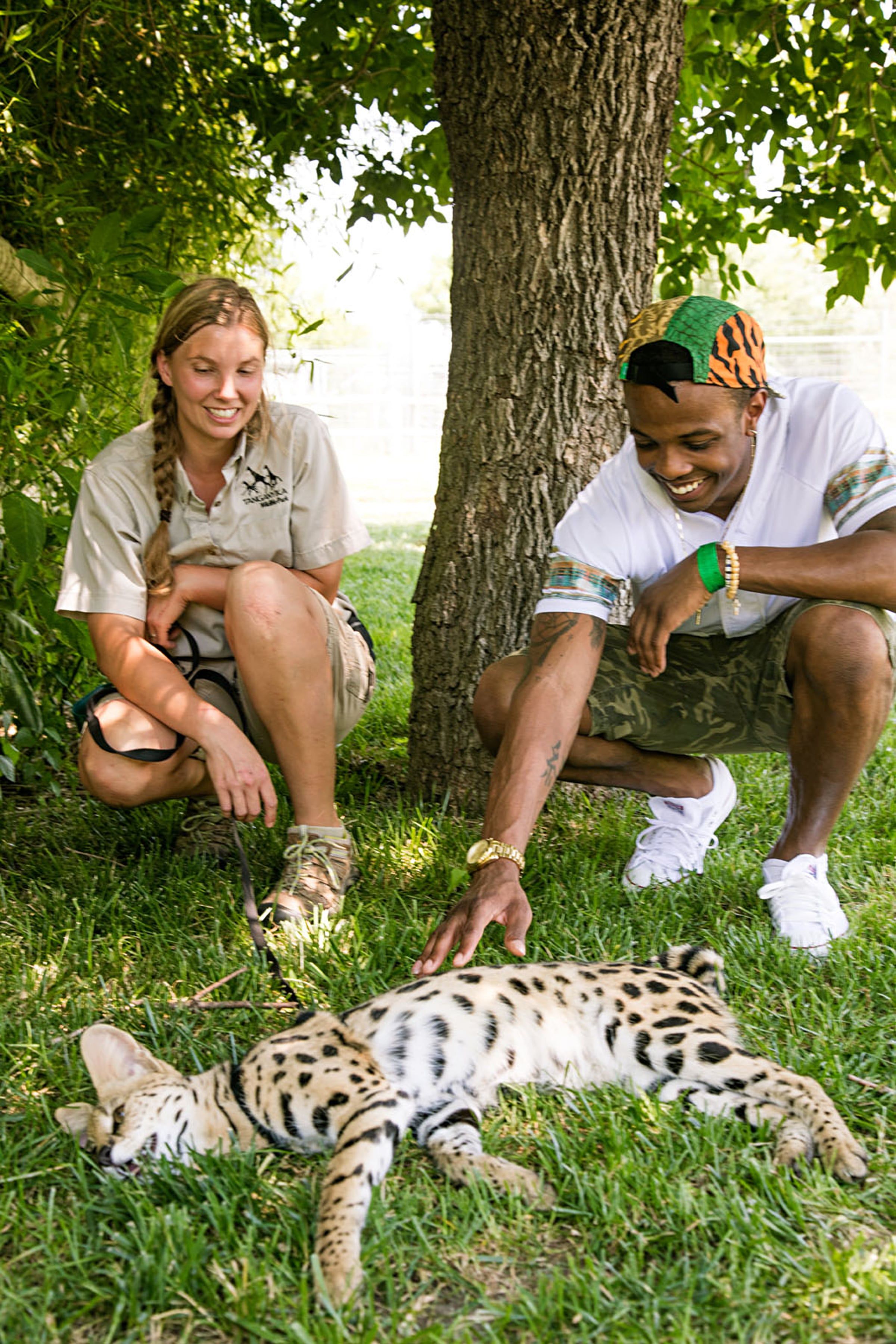 A Tanganyika Wildlife Park staff member and a visitor crouching in the grass beside a serval