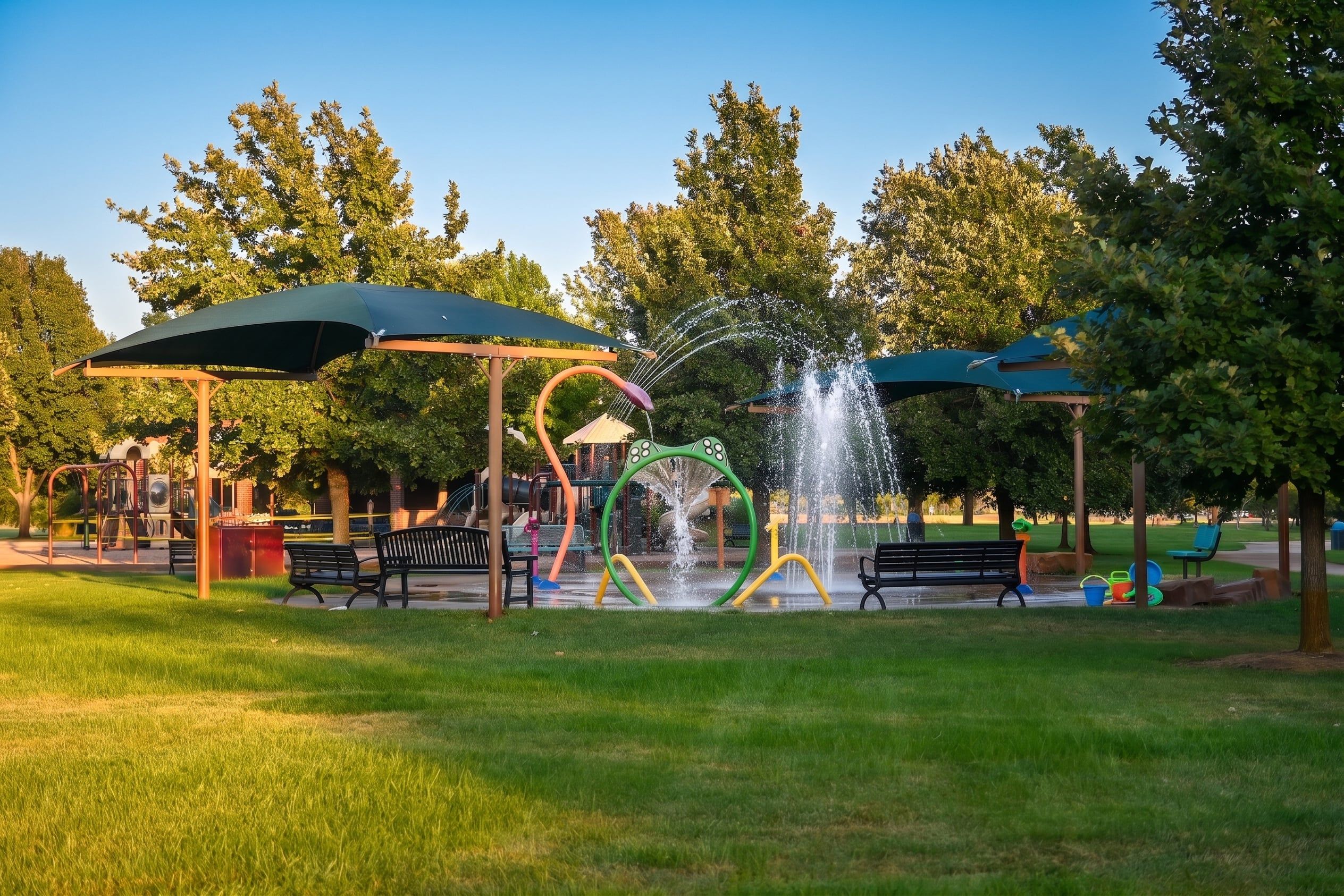 The McLaughlin Park splash pad with its water features running, surrounded by shaded benches, playground equipment, and mature trees