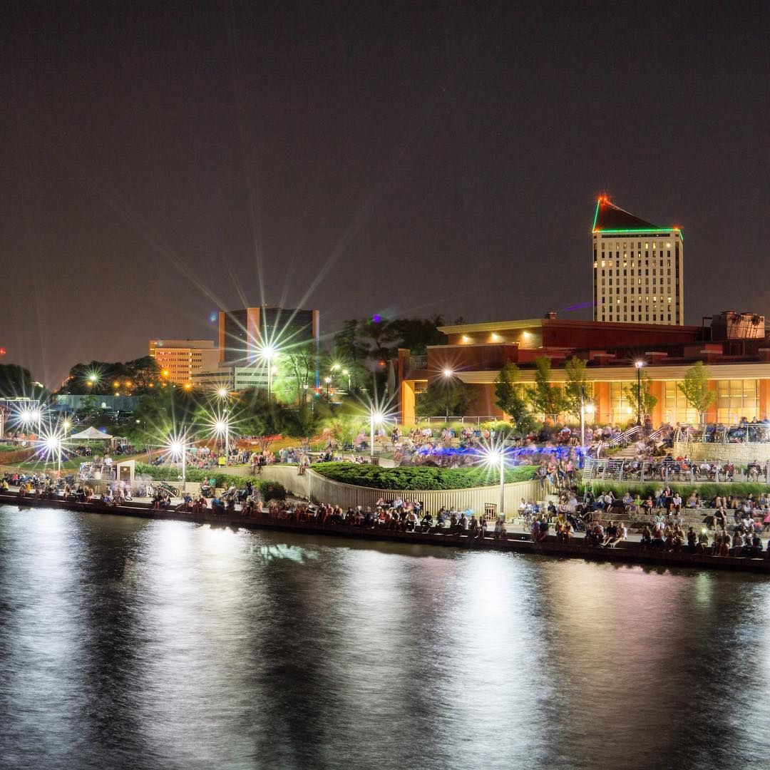 Crowds lining the bank of the Arkansas River at night during Wichita Riverfest, with the lit downtown skyline behind them